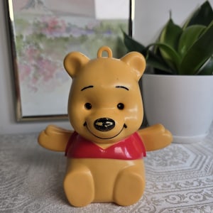May include: A yellow plastic Winnie the Pooh figurine featuring a red shirt, black painted eyes, eyebrows, and a mouth. The bear has a small loop at the top of its head, outstretched arms, and a worn black paint finish on the nose area.