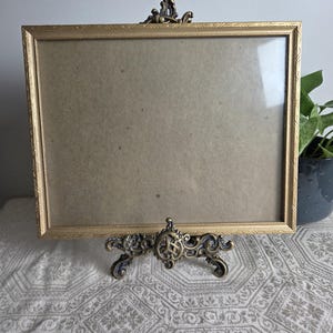 May include: A rectangular gold picture frame with a textured border, supported by an ornate, dark bronze-toned metal easel stand featuring intricate scrollwork and a central circular emblem, holding a plain, light brown cardboard backing.
