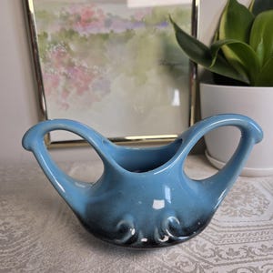 May include: A light blue ceramic vase with two curved handles and a dark blackish-blue gradient at the base. The vase features a wide, open top and a rounded body with decorative moulded swirls near the bottom, showcasing a smooth, glossy finish.