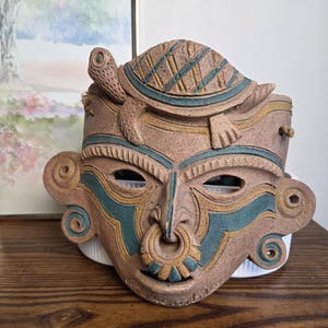 May include: A handcrafted ceramic mask with a turtle design on top. The mask features a face with a nose ring and teal and yellow accents. The mask is brown and tan in color and has a unique, artistic design.