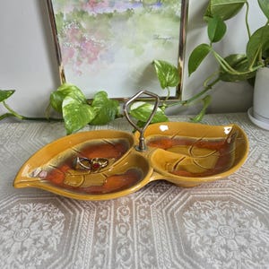 Puede incluir: Un plato de servicio de cerámica de dos compartimentos con forma de dos hojas conectadas y un asa central de metal. El plato presenta un esmalte brillante en tonos amarillo mostaza, naranja quemado y marrón, con dos anillos dentro de un lado.