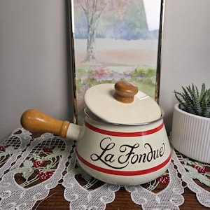May include: A cream-coloured enamel fondue pot with a speckled finish, featuring a wooden handle and a matching lid with a wooden knob. The pot is decorated with two horizontal red stripes and the black cursive text "La Fondue" on the front.