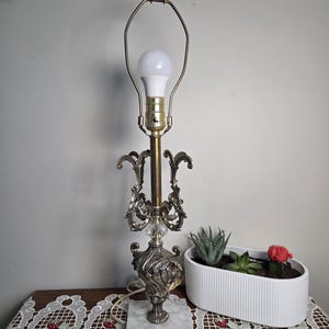 May include: An ornate brass and silver table lamp with a white bulb. The lamp has a decorative base and a marble square base. A white planter with succulents and a red cactus sits nearby on a table with a lace runner.