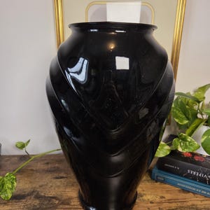 Puede incluir: Un jarrón grande de color negro brillante con una abertura ancha y redondeada y una base cónica. La superficie exterior presenta crestas prominentes en forma de V que envuelven el cuerpo, creando una apariencia texturizada y superpuesta.