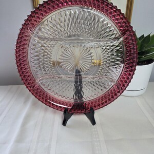 May include: A round, clear glass serving platter featuring a deep magenta-coloured rim with a textured diamond pattern. The centre of the platter is divided into three sections by raised glass dividers and displays a sunburst pattern in the middle.
