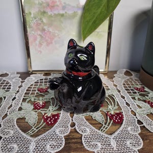 May include: A black cat ornament with green eyes and a red collar. The cat is positioned on a white lace doily with strawberry embroidery. The ornament is a decorative item.