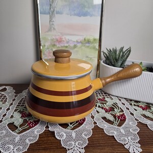 Peut inclure: Une casserole en émail jaune avec un couvercle, ornée de rayures horizontales dans des tons de brun et de brun foncé. La casserole possède un manche en bois et un bouton en bois assorti sur le couvercle. Le bord est fini en métal argenté.