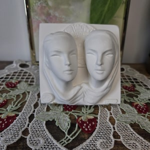 May include: A white rectangular wall plaque featuring a relief sculpture of two faces side by side. The faces are smooth and stylised with draped fabric elements around the necks. A circular zodiac-style emblem is visible in the background.