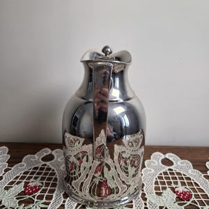 Puede incluir: Una jarra de color plateado con asa y un dise&ntilde;o decorativo de fresas. La jarra tiene una superficie reflectante y una tapa. El dise&ntilde;o de fresas est&aacute; en la parte inferior de la jarra.