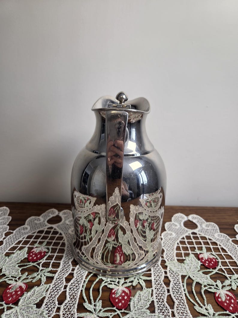 Puede incluir: Una jarra de color plateado con asa y un dise&ntilde;o decorativo de fresas. La jarra tiene una superficie reflectante y una tapa. El dise&ntilde;o de fresas est&aacute; en la parte inferior de la jarra.