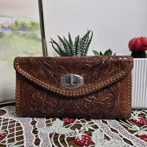 May include: A brown leather clutch bag with a floral embossed pattern and a silver clasp. The bag has a stitched border and a flap closure. The background includes a framed watercolour painting and a white planter with succulents.