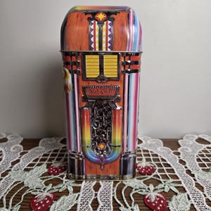 Può includere: Una scatola di latta rettangolare progettata per assomigliare a un jukebox vintage. La scatola presenta un design colorato con le parole "Rock & Roll!!" e un motivo a stella. I lati hanno strisce verticali di vari colori.