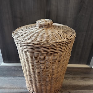May include: A tall, cylindrical woven wicker hamper featuring a natural light brown finish. The basket includes a matching circular lid with a small, centred knob handle on top. The structure is crafted from tightly woven reeds in a vertical pattern.