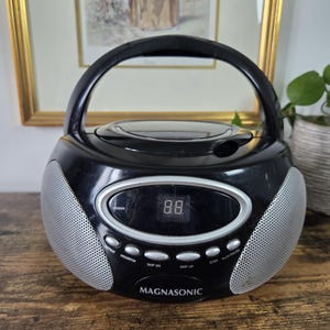 以下が含まれることがあります： 丸みを帯びたデザインの黒いMagnasonic製ポータブルCDプレーヤー。上部に持ち運び用のハンドルがあり、両サイドに銀色のメッシュスピーカーグリルを備えています。前面には「88」と表示されたデジタルディスプレイ、操作ボタン、ブランド名が配置されています。