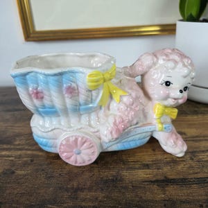 Puede incluir: Una maceta de cerámica con forma de cordero rosa tirando de un cochecito de bebé azul y blanco. El cochecito tiene un lazo amarillo, mientras que el cordero lleva una pajarita amarilla. Las ruedas son rosas con diseño floral y tiene flores rosas.