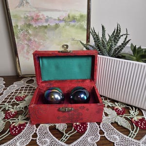 May include: A small, open red box with a distressed finish, lined with green fabric, contains two iridescent spheres. The box is on a table with a white lace runner featuring strawberry embroidery. A framed watercolour painting and a white planter with succulents are in the background.