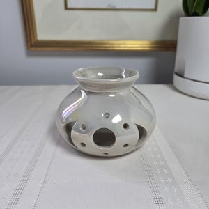 May include: A round, pearlescent white ceramic oil burner with a wide opening at the top and a series of circular cutouts around the body. The surface has a glossy, iridescent finish that reflects light, and the structure features a central larger hole surrounded by smaller, evenly spaced circular perforations.