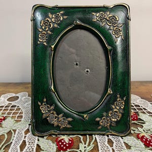 Puede incluir: Un marco de fotos verde oscuro con una abertura ovalada. El marco tiene adornos florales dorados en cada esquina y a lo largo de los bordes. El marco está sobre un mantel individual de encaje blanco con bordados de fresas.