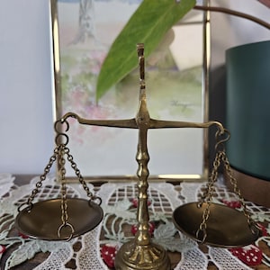 Puede incluir: Una balanza de latón vintage con dos platillos redondos suspendidos por cadenas. La balanza tiene una base decorativa y una columna central. El fondo incluye un cuadro enmarcado y una hoja verde.