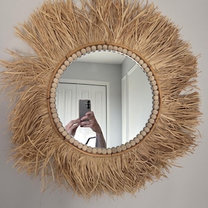 May include: A round wall mirror featuring a central glass pane surrounded by a frame of light-colored wooden beads. The outer edge is decorated with a thick, textured fringe made of natural tan raffia fibers that extend outward in a sunburst pattern.