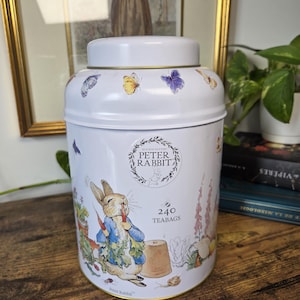 以下が含まれることがあります： 「Beatrix Potter Peter Rabbit」および「240 teabags」という文字が前面に印刷された、ピーターラビットのイラスト入り白い円筒形の金属製ティー缶。缶にはカラフルな蝶や庭をテーマにした絵が描かれています。