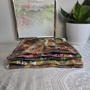 May include: A stack of four square quilted fabric coasters featuring various autumn-inspired patterns in shades of brown, mustard yellow, olive green, and deep red. Each coaster has a stitched diamond pattern and finished edges with visible fabric textures.