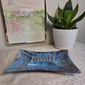 Puede incluir: Un cenicero rectangular de cerámica con un esmalte brillante en tonos azul, morado y marrón. La pieza tiene una forma cóncava distintiva con cuatro apoyos para cigarrillos integrados a lo largo de la cresta central y un acabado moteado.