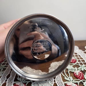 Puede incluir: Primer plano de la base de un termo Stanley plateado. La base circular tiene el texto "STANLEY IT WILL NOT BREAK" y "LANDERS, FRIARY & CLARK NEW BRITAIN, CONN. U.S.A.". Tambi&eacute;n es visible el n&uacute;mero de patente D:098.