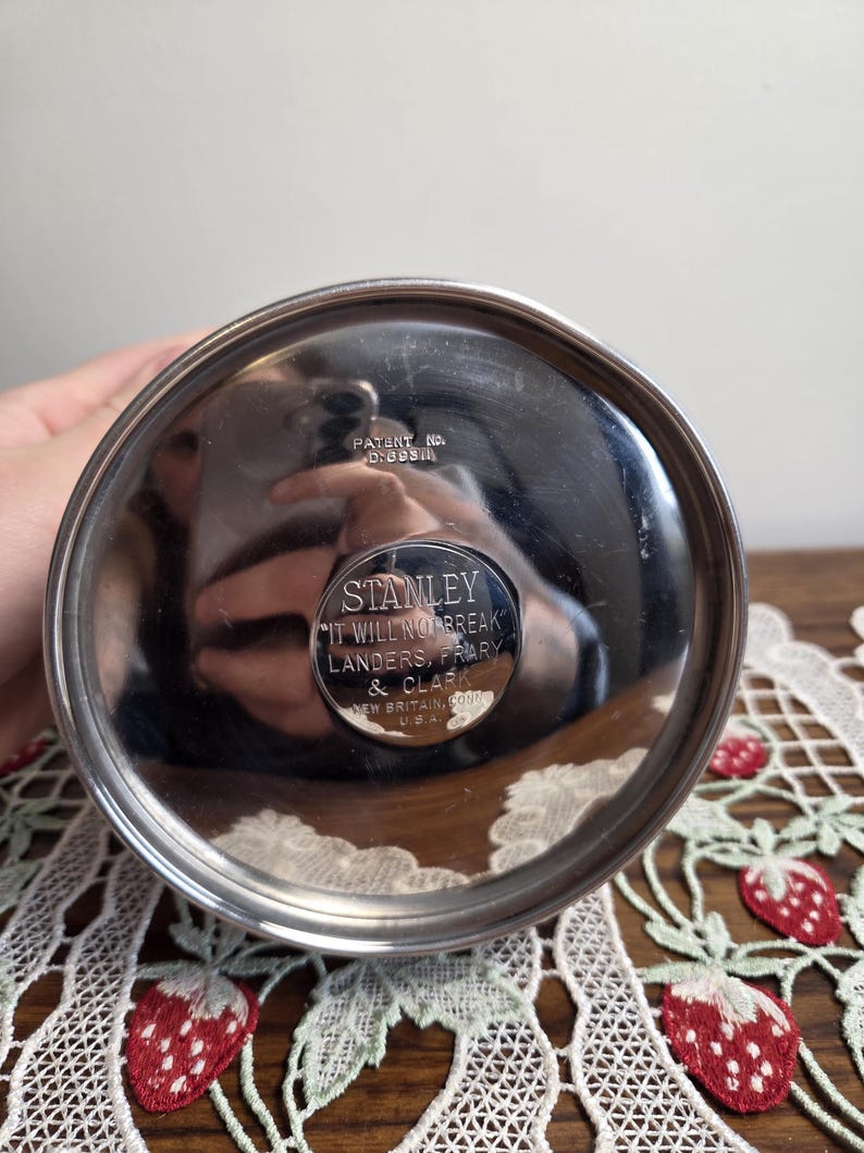Puede incluir: Primer plano de la base de un termo Stanley plateado. La base circular tiene el texto "STANLEY IT WILL NOT BREAK" y "LANDERS, FRIARY & CLARK NEW BRITAIN, CONN. U.S.A.". Tambi&eacute;n es visible el n&uacute;mero de patente D:098.