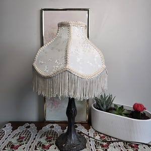 Könnte beinhalten: Eine antike Tischlampe mit einem weißen, floral gemusterten Lampenschirm, der mit cremefarbenen Fransen und Zierleisten versehen ist. Die Lampe hat einen dunklen Metallsockel und steht auf einer Holzoberfläche mit einem Spitzentischläufer. Ein gerahmtes Bild und ein weißer Pflanztopf mit Sukkulenten befinden sich im Hintergrund.
