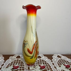 May include: A tall, decorative glass vase with a flared, ruffled rim. The vase features a vibrant color scheme, transitioning from a red rim to yellow, and a creamy white body with swirling orange, green, and brown accents. The vase sits on a clear, circular base.
