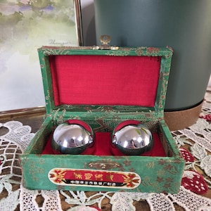 May include: A vintage green box with a red interior holds two shiny, silver metal spheres. The box has a gold clasp and a decorative label with Chinese characters. The box is on a lace tablecloth.
