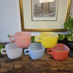 May include: A set of five square-shaped mugs in various pastel colours including white, pink, yellow, light blue, and coral. Each mug features a small, square handle on the side and a smooth, opaque finish. The mugs are arranged in a cluster.