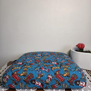 May include: A square, blue pillow with a repeating pattern of colorful cowboy boots. The boots are red, brown, white, and blue, with various designs. The pillow is on a wooden surface with a white lace trim.