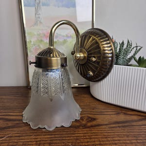 May include: A vintage wall sconce with a brass finish. The sconce has a frosted glass shade with a scalloped edge and a decorative pattern. The fixture is mounted on a brass backplate and has a curved arm. It is designed for indoor lighting.