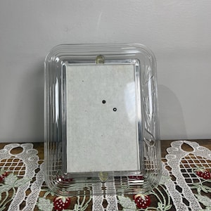 Puede incluir: Un marco de fotos rectangular transparente con un borde acanalado. El marco tiene un pequeño elemento decorativo en la parte superior y está diseñado para contener una fotografía. El marco está sobre una mesa con un mantel individual de encaje con tema de fresas.