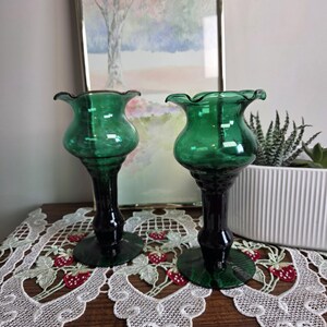 Peut inclure: Deux bougeoirs en verre vert avec des bords festonnés et des tiges vert foncé. Les bougeoirs sont posés sur une surface en bois avec un chemin de table en dentelle blanche avec des fraises brodées. Une peinture encadrée est en arrière-plan.