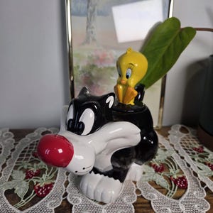 May include: A ceramic figurine of Sylvester the Cat with Tweety Bird perched on top. Sylvester is black and white with a red nose. Tweety is yellow with blue eyes. The figurine is on a wooden surface.