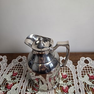 Puede incluir: Una jarra plateada con tapa y asa. La jarra tiene un cuerpo redondeado y un pico. La parte inferior de la jarra tiene un dise&ntilde;o decorativo. La jarra est&aacute; sobre un mantel de encaje blanco con dise&ntilde;os de fresas rojas.