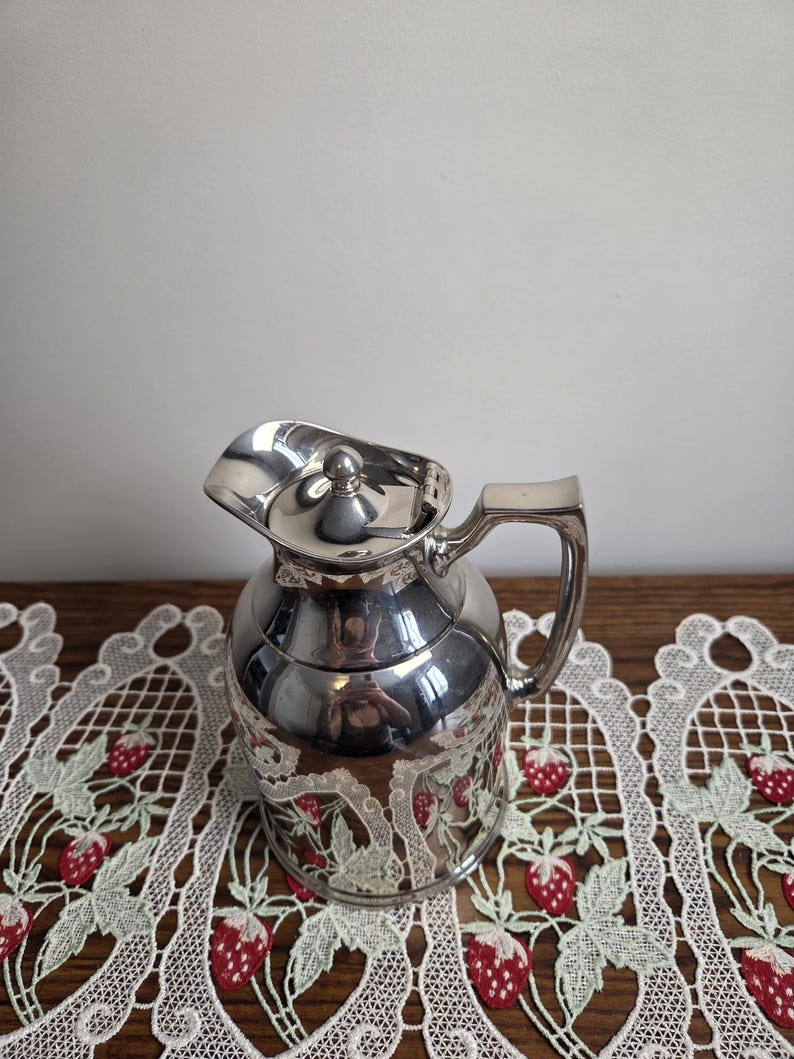 Puede incluir: Una jarra plateada con tapa y asa. La jarra tiene un cuerpo redondeado y un pico. La parte inferior de la jarra tiene un dise&ntilde;o decorativo. La jarra est&aacute; sobre un mantel de encaje blanco con dise&ntilde;os de fresas rojas.