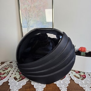May include: A spherical vase featuring a glossy black upper section and a matte black lower section with three distinct horizontal ridges. The vase has a circular opening at the top and a smooth, rounded body that transitions between textures.