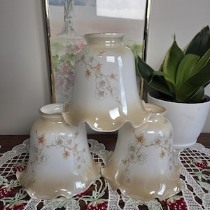 Peut inclure: Un ensemble de trois abat-jours en verre en forme de cloche, dotés d'une base blanche avec un bord dégradé beige et un motif délicat de branches fleuries brunes. Chaque abat-jour présente un bord ondulé et une ouverture circulaire au sommet.