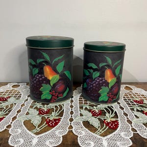 May include: Two metal canisters with lids. The canisters are black with a painted design of fruit, including pears, grapes, and plums. The lids are green. The canisters are on a wooden surface with a white lace runner.