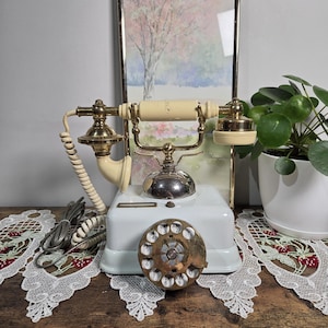 May include: Vintage white rotary phone with brass accents. The phone features a cream-colored handset and a dial with numbers. The phone is displayed on a wooden surface with a decorative lace runner and a framed picture in the background.