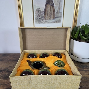 Puede incluir: Un juego de té en una caja de cartón color canela con un forro de satén naranja brillante. El set incluye una tetera marrón oscuro y seis tazas de té pequeñas a juego, todas con un esmalte verde moteado en los bordes superiores y las tapas.