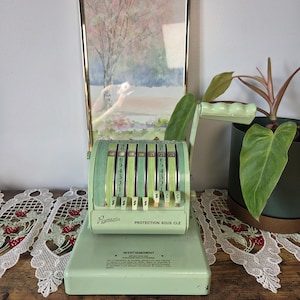 May include: A vintage light green Paymaster check writer machine featuring a manual hand crank, vertical sliding number levers, and the text "Paymaster" and "PROTECTION SOUS CLE" printed on the front, along with a warning label on the base.