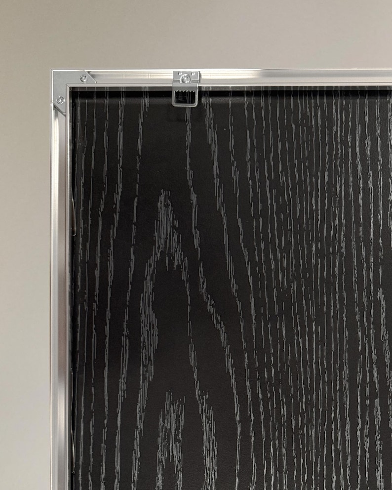 Puede incluir: Primer plano de un panel enmarcado con vetas de madera en negro y gris. El marco es de metal plateado. Un colgador de metal est&aacute; fijado en la parte superior del marco.