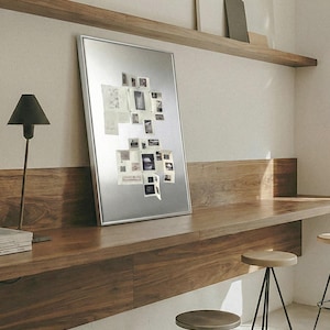 Puede incluir: Un tablero magn&eacute;tico con marco plateado que muestra varias fotos y notas peque&ntilde;as. El tablero est&aacute; apoyado contra un escritorio de madera, con una l&aacute;mpara negra y una pila de libros a la izquierda. La habitaci&oacute;n tiene un dise&ntilde;o minimalista.