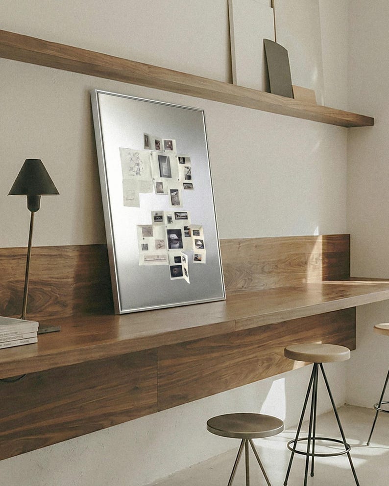 Puede incluir: Un tablero magn&eacute;tico con marco plateado que muestra varias fotos y notas peque&ntilde;as. El tablero est&aacute; apoyado contra un escritorio de madera, con una l&aacute;mpara negra y una pila de libros a la izquierda. La habitaci&oacute;n tiene un dise&ntilde;o minimalista.