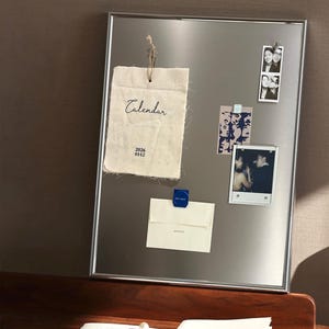 Puede incluir: Un tablero magn&eacute;tico con marco plateado que muestra un calendario de tela, fotos y notas. El calendario es de tela texturizada blanquecina con la palabra "Calendar" y la fecha "2026 01-12" impresas. Otros elementos se adjuntan con imanes.